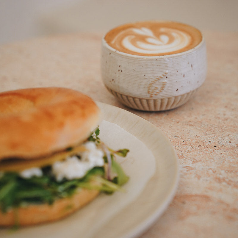 Cappuccino mit Latte Art in einer unbound-Kaffeetasse und ein belegter   Bagel auf einem Teller